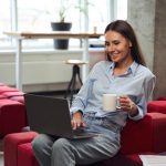 Cheerful young woman with teacup in hand looking at computer monitor while sitting on chair in comfortable coworking space