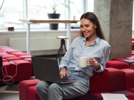 Cheerful young woman with teacup in hand looking at computer monitor while sitting on chair in comfortable coworking space