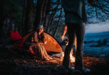 Ouverture 11 2026, Argentine | Huttopia plante ses tentes à Mar Azul A happy camper sitting by a warm campfire near a tent and chair beside a tranquil lake as night falls.