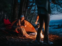 A happy camper sitting by a warm campfire near a tent and chair beside a tranquil lake as night falls.