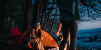 Ouverture 11 2026, Argentine | Huttopia plante ses tentes à Mar Azul A happy camper sitting by a warm campfire near a tent and chair beside a tranquil lake as night falls.