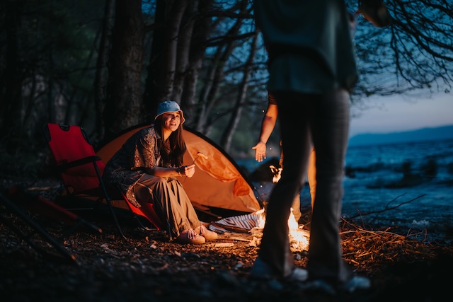 A happy camper sitting by a warm campfire near a tent and chair beside a tranquil lake as night falls.